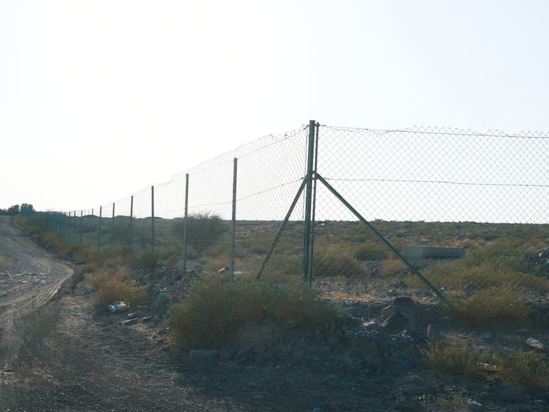 A chain-link fence along a dirt road with sparse vegetation and a bright sky in the background.