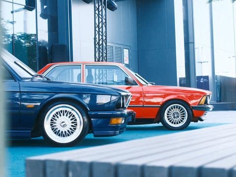 Two classic cars, a blue BMW and a red BMW, parked side by side in a modern urban setting.