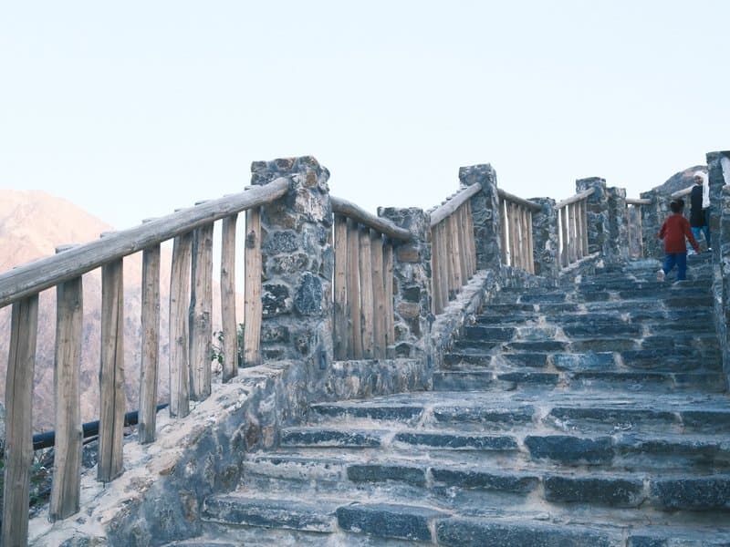 A person walking up stone steps with wooden railings, surrounded by rocky terrain and mountains.