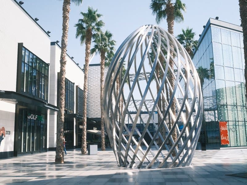 A modern metallic sculpture resembling an egg, surrounded by palm trees and contemporary buildings.