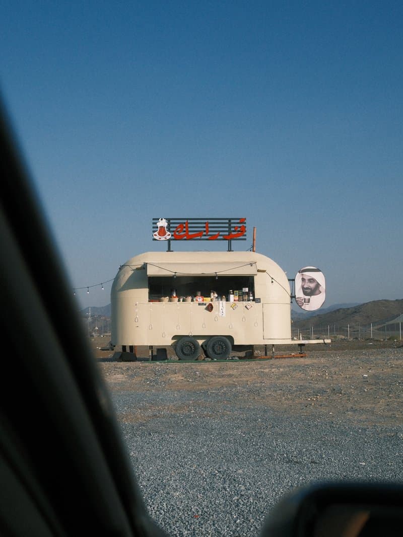 A food truck with a large sign, set against a clear blue sky and gravel ground.