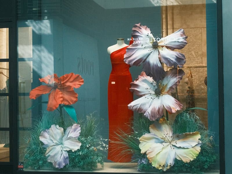 A vibrant window display featuring large flowers and a red dress in a boutique setting.