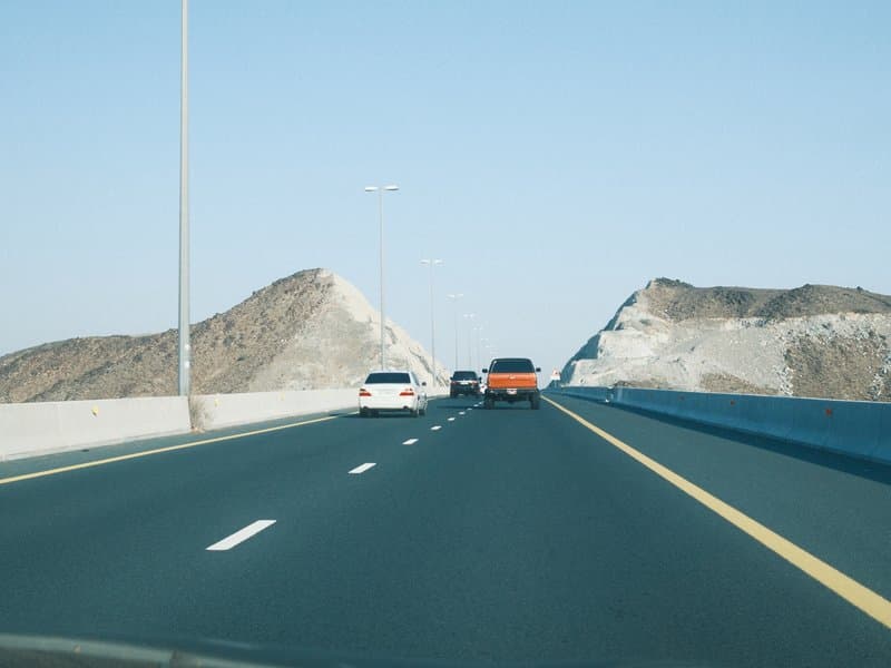 A highway scene with two cars driving towards rocky hills under a clear blue sky.