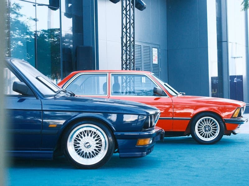 Two classic cars, a blue convertible and a red sedan, parked side by side in an urban setting.